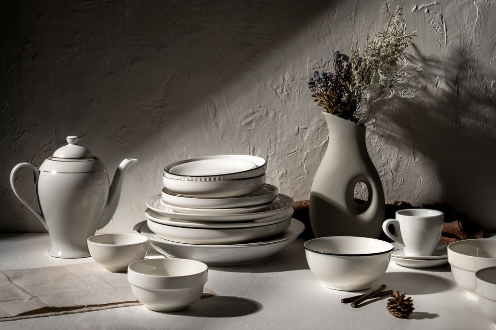 Elegant white ceramic dinnerware and teapots arranged in dramatic lighting against textured wall.