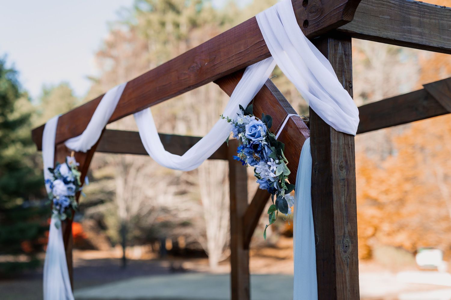 Wedding ceremony arch decorated with white fabric draping and blue flower accents in outdoor setting.