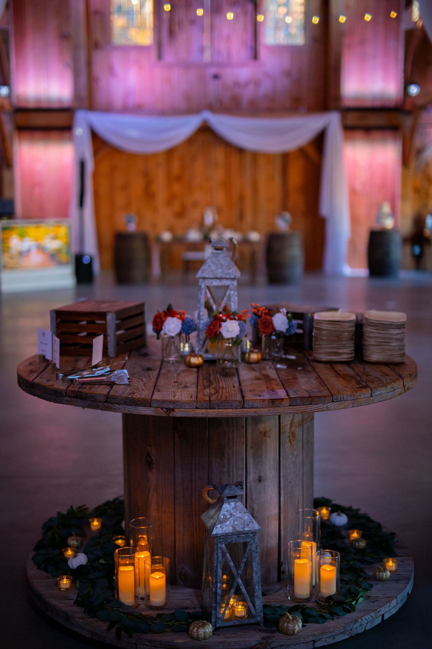 Rustic wedding decor featuring wooden spool table with lanterns and candles against pink-lit wooden backdrop.