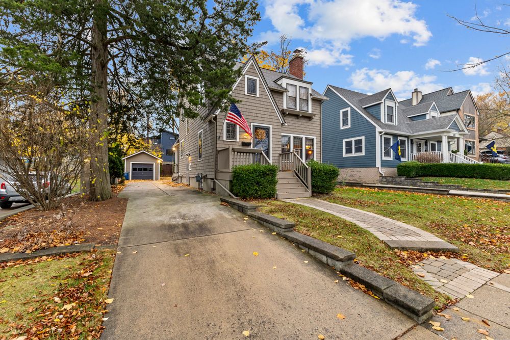 Front exterior of a gray Cape Cod home with a brick walkway, detached garage, and autumn landscaping.