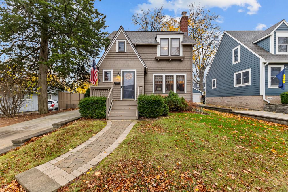 Charming gray Cape Cod home with an American flag, brick pathway, green lawn, and colorful fall foliage.