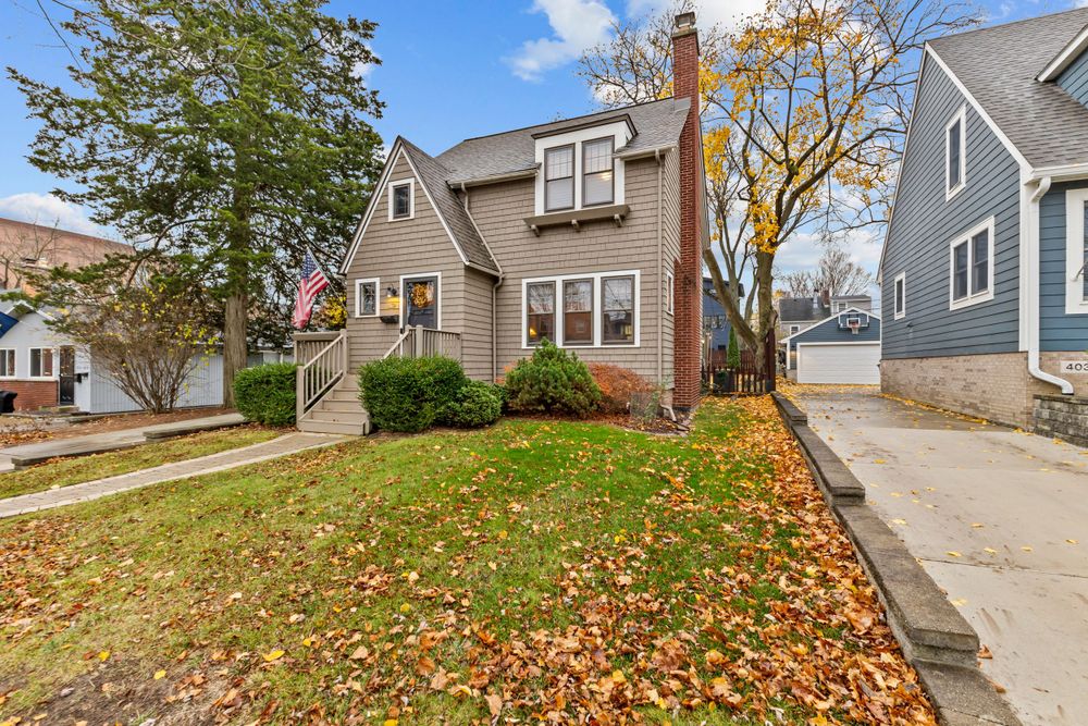 Gray Cape Cod style home with brick chimney, green lawn, and fall leaves along a residential street.