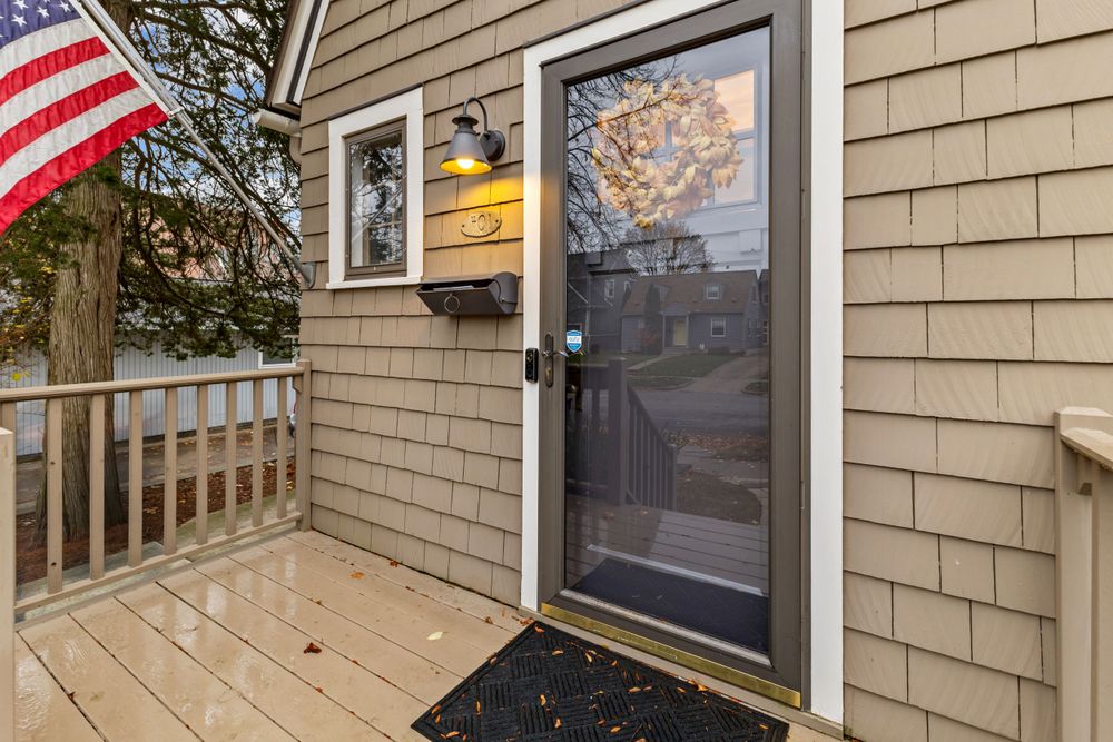 Close-up of a gray shingle home entry with a glass storm door, wall lantern, and wooden deck.