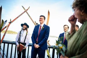 Groom in navy suit smiling during outdoor lakeside wedding ceremony framed by vintage crossed skis.