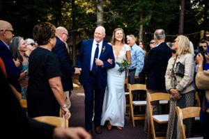 Bride in white gown walks down outdoor aisle with escort among seated wedding guests in the woods.