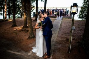 Newlyweds share a romantic first kiss near a lantern with wedding guests gathered by the lake.