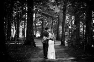 Black and white photo of bride and groom kissing on a forested path surrounded by tall trees.