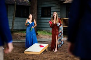 Two women in formal dresses laughing while playing cornhole at an outdoor wedding reception.