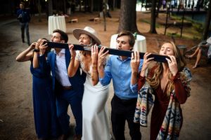 Wedding guests playfully drinking from long black tubes at an outdoor forest reception.