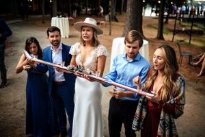 Bride in white dress and hat holds a large novelty item with wedding guests outdoors.