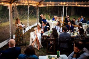Wedding guest delivers a heartfelt speech to guests seated under a clear outdoor tent.