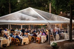 Large wedding reception under a clear tent in a wooded outdoor setting at dusk.