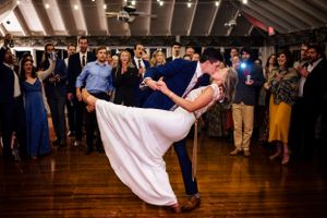 Bride and groom share a romantic dip kiss on the dance floor surrounded by cheering wedding guests.