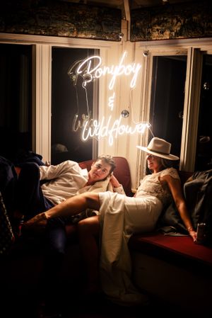 Newlyweds relax together beneath a glowing neon sign reading Ponyboy and Wildflower at their wedding.