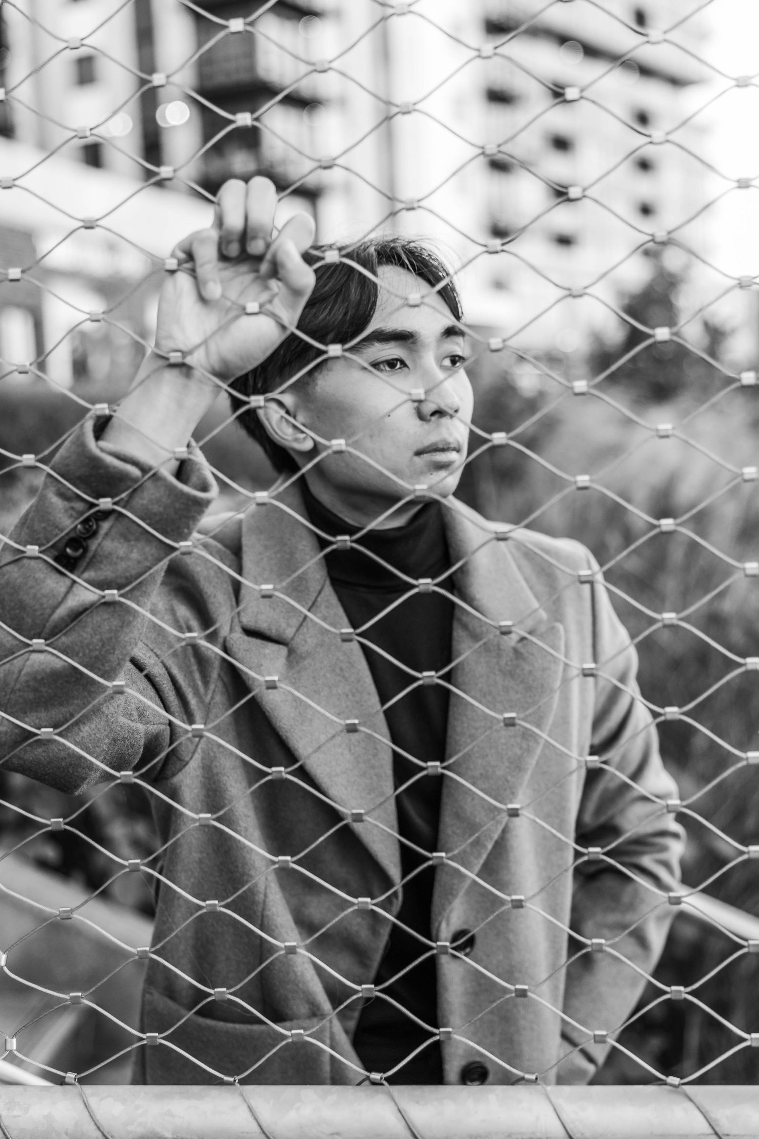 Young Asian man in camel coat standing behind chain-link fence in black and white urban photo.