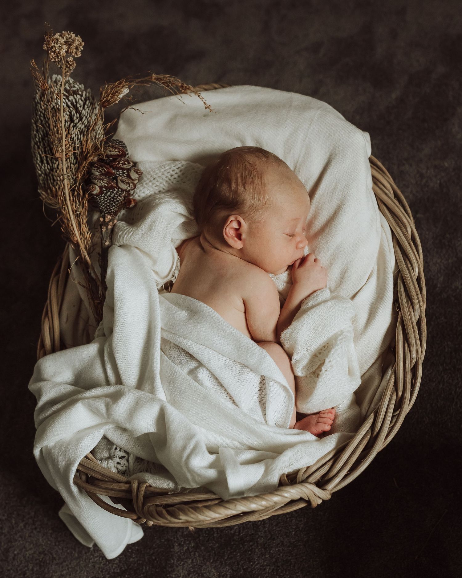 Sleeping newborn baby wrapped in soft white blankets in a woven basket with dried flowers.
