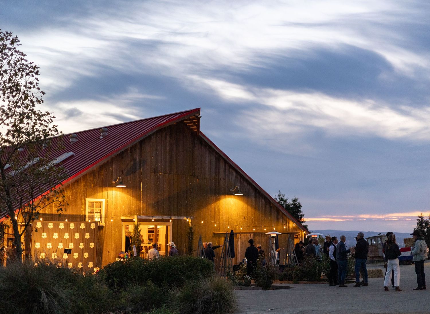 A rustic barn wedding venue illuminated at dusk with guests gathered outside under warm glowing lights.