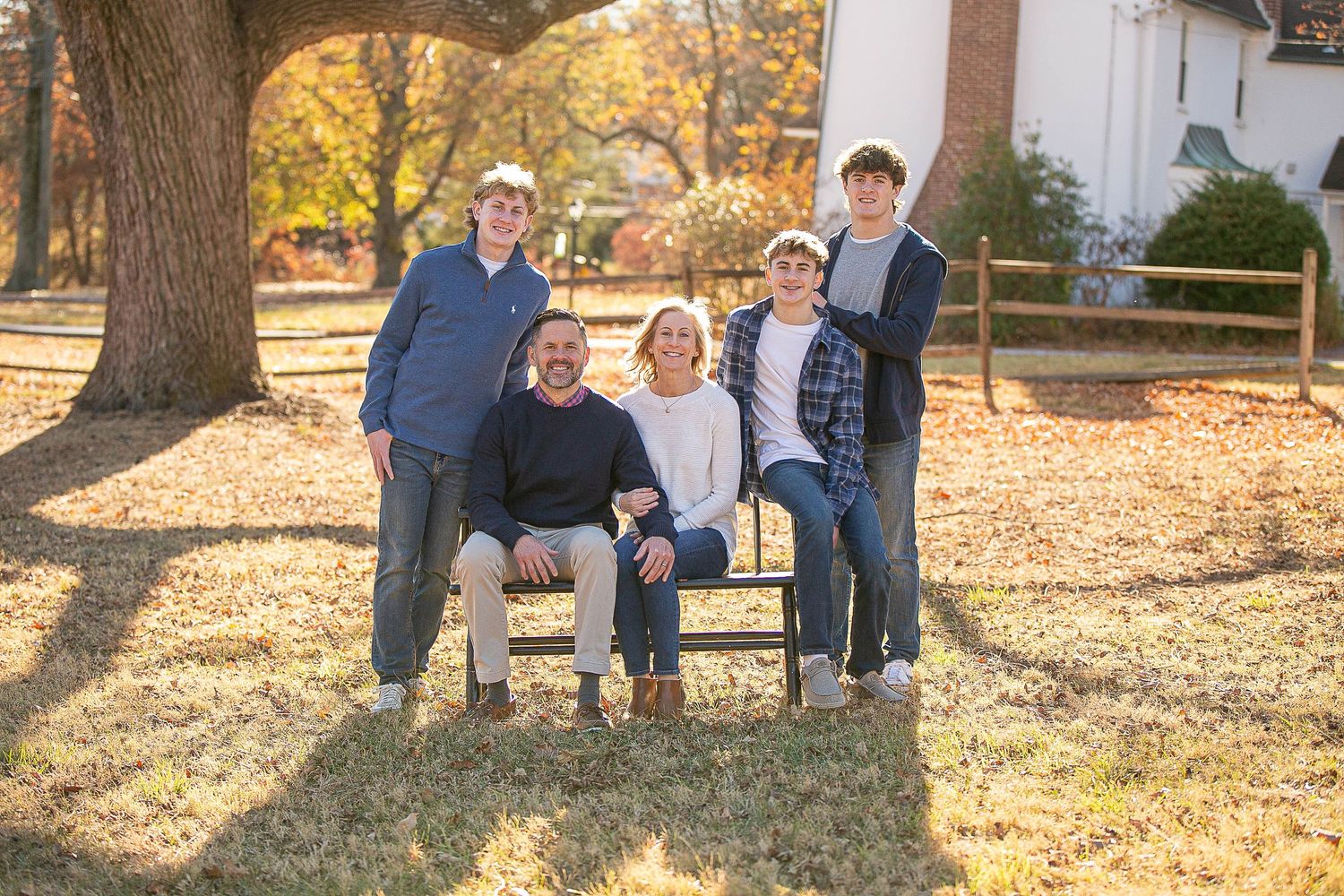 Family group seated on a wooden bench under a large tree with fallen autumn leaves.