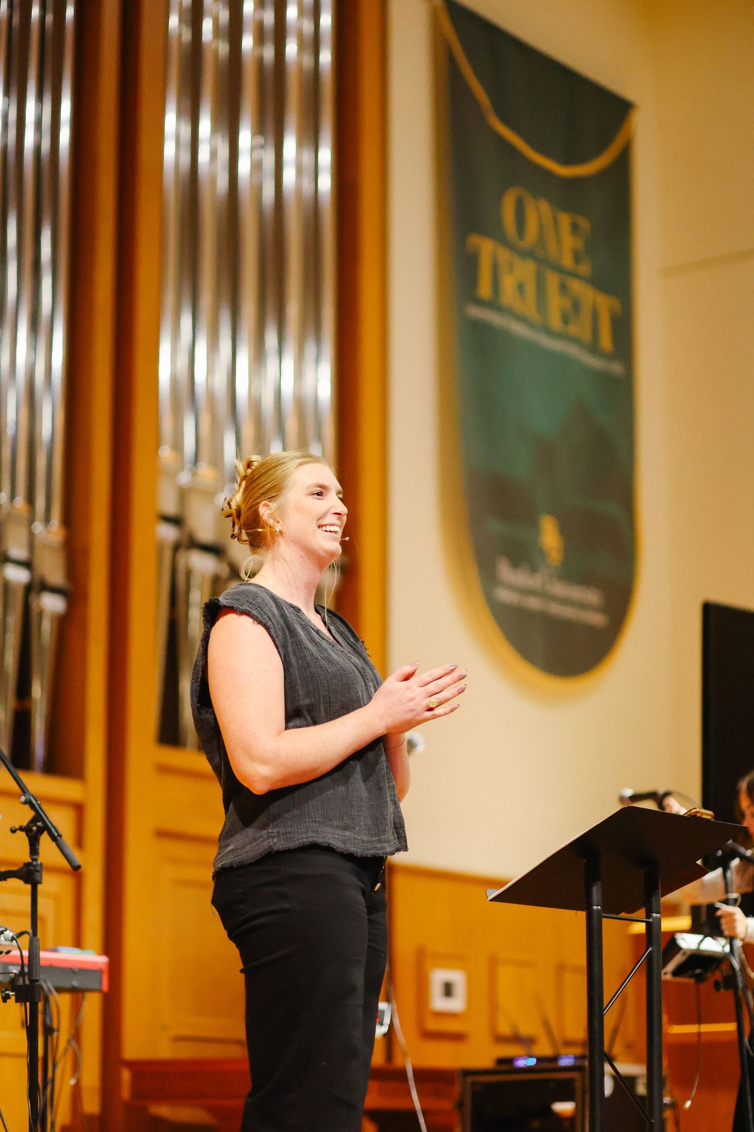 A woman speaking and clapping on stage in a church with a large pipe organ behind her.