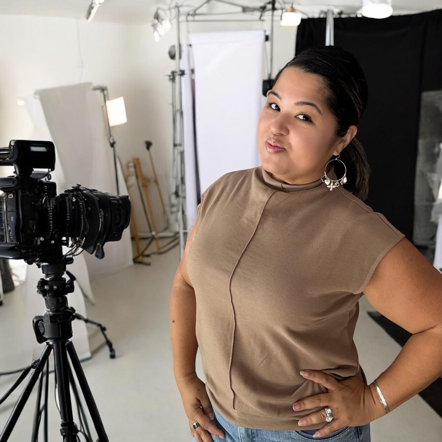 Photography studio setup with camera equipment and person in beige top posing beside tripod.