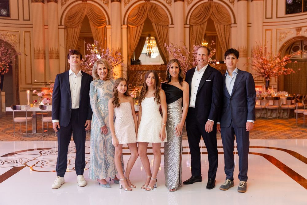 Formal group portrait in elegant dresses and suits standing in an ornate ballroom.