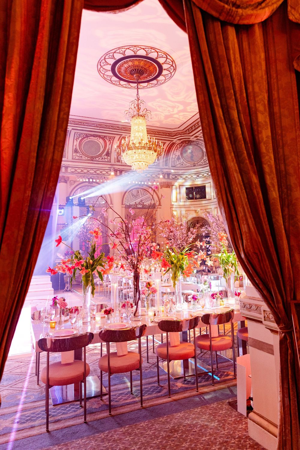 Opulent ballroom setup with pink lighting, crystal chandeliers and tall floral centerpieces on dining tables.