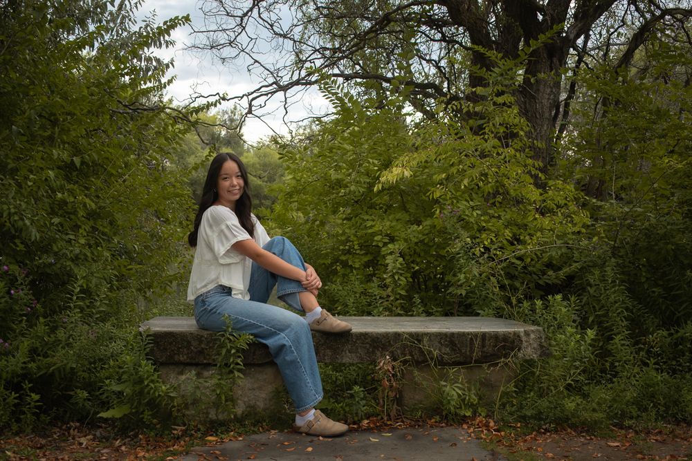 A figure sits thoughtfully on a wooden bench surrounded by lush green foliage in a peaceful garden setting.