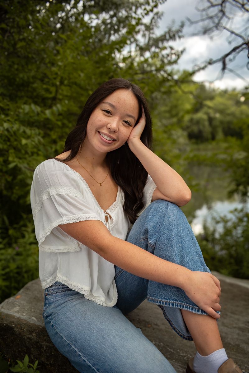 A casual outdoor portrait session featuring someone in a white blouse and blue jeans sitting on rocks near trees.