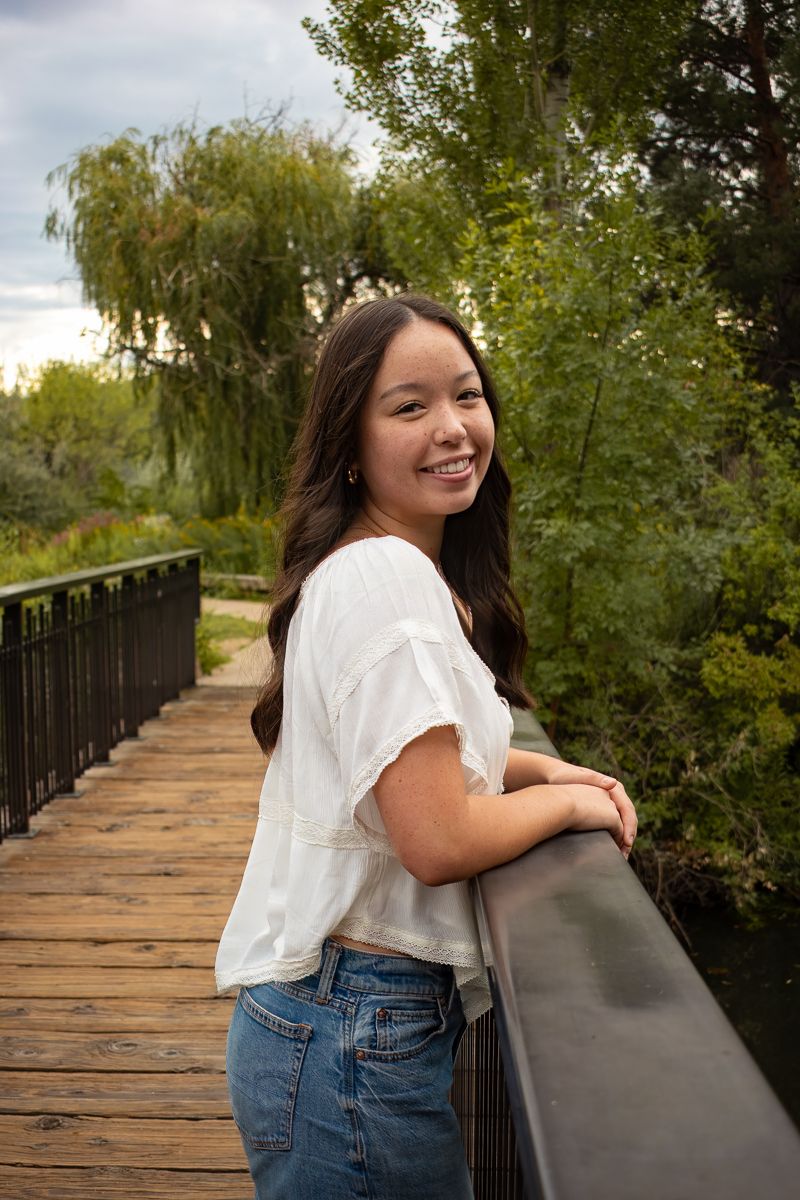 A person in a white shirt and jeans leans against a wooden bridge railing surrounded by lush green trees.