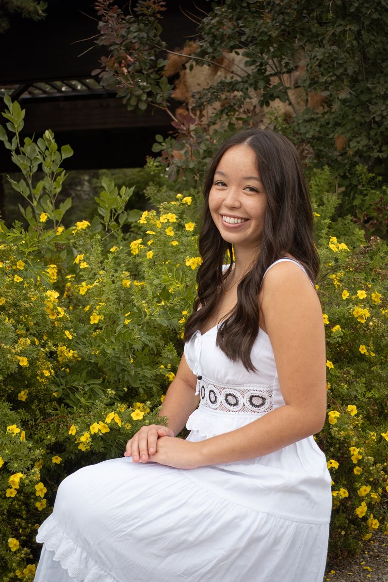 A person in a white dress sits outdoors against a backdrop of yellow flowers and green foliage in a garden setting.
