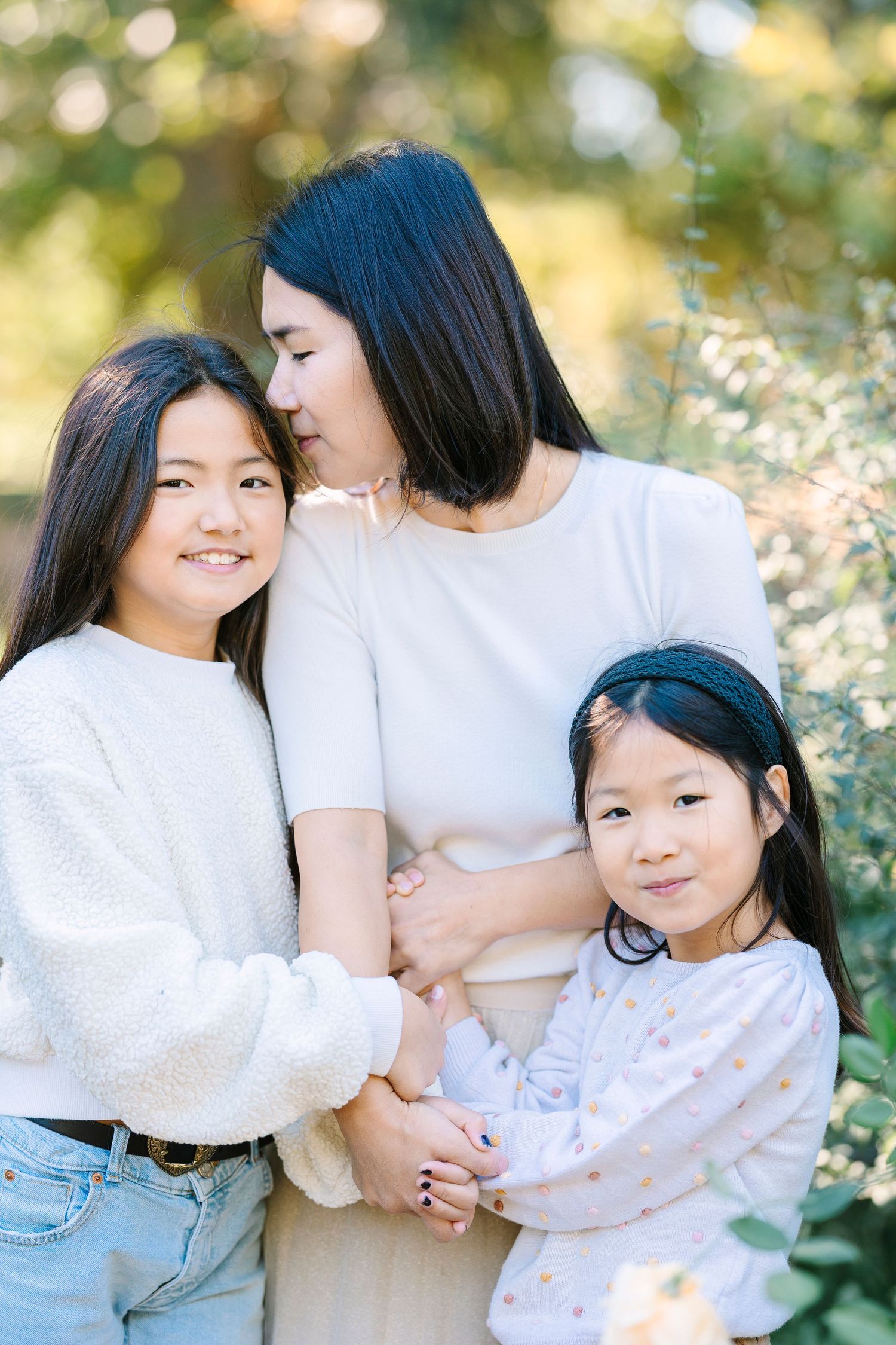 Three people in white tops share tender moments together outdoors with soft bokeh lighting in the background.