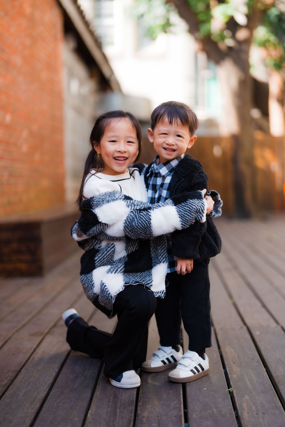 A sister hugs her younger brother while kneeling on an outdoor wooden patio