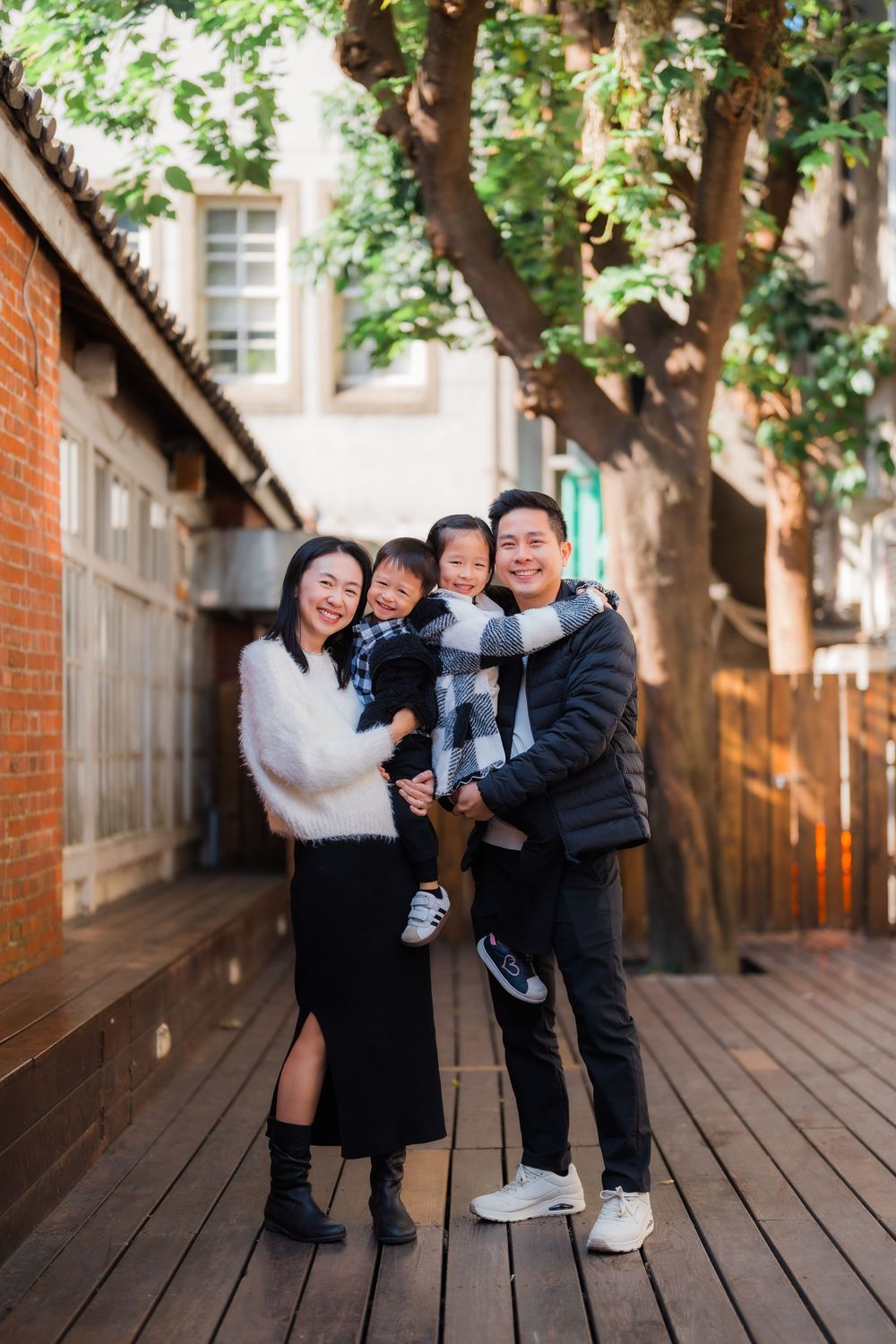 A family of four pose on a wooden deck with brick walls and greenery for a portrait in Taipei's Huashan 1914 Creative Park.