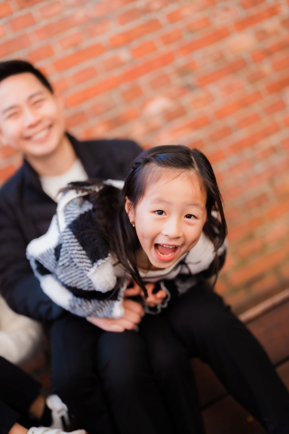 A father playfully swings his daughter around in front of a brick wall at Huashan Creative Park