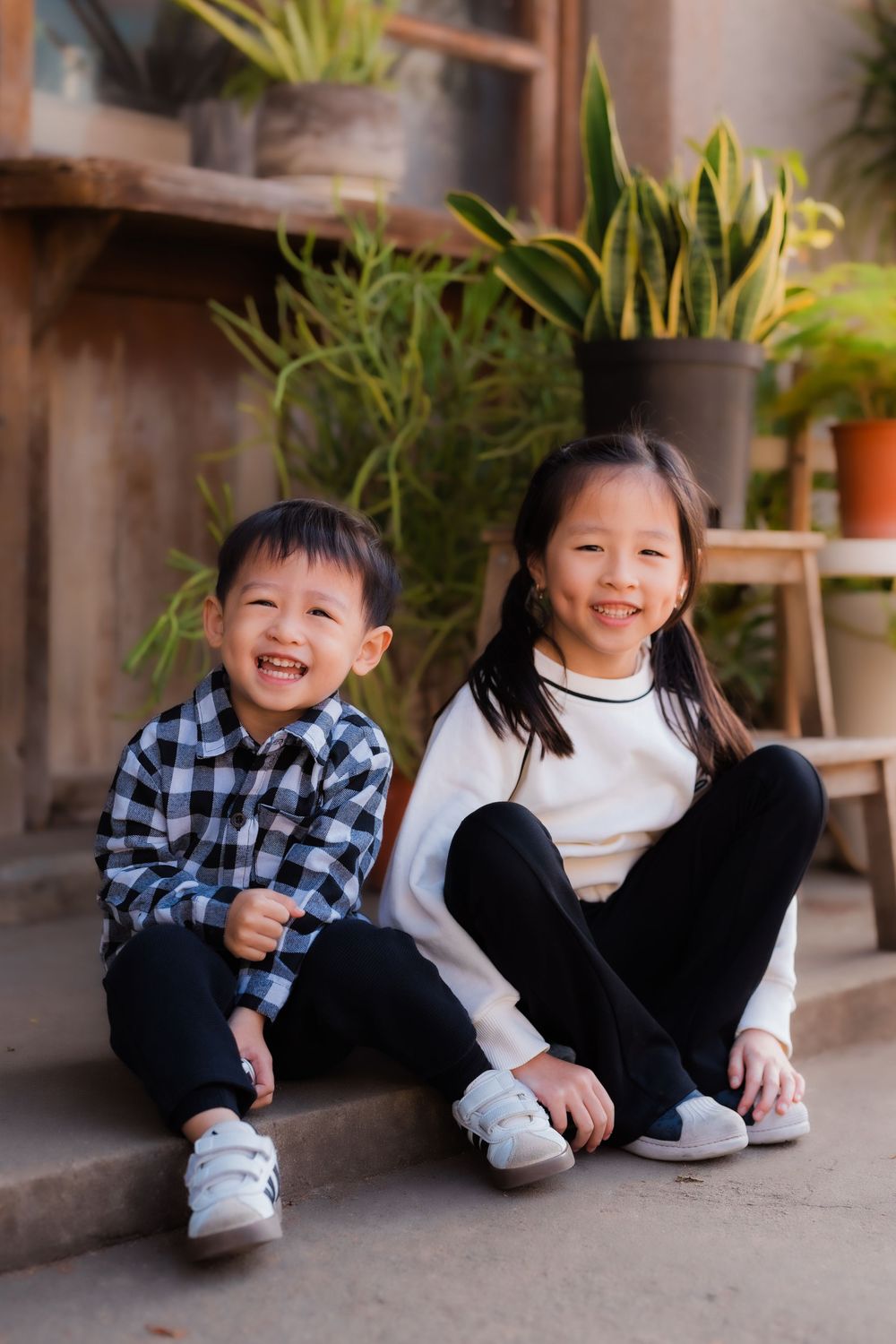 Two children laughing together sitting on a patio with potted plants and wooden accent features in background.