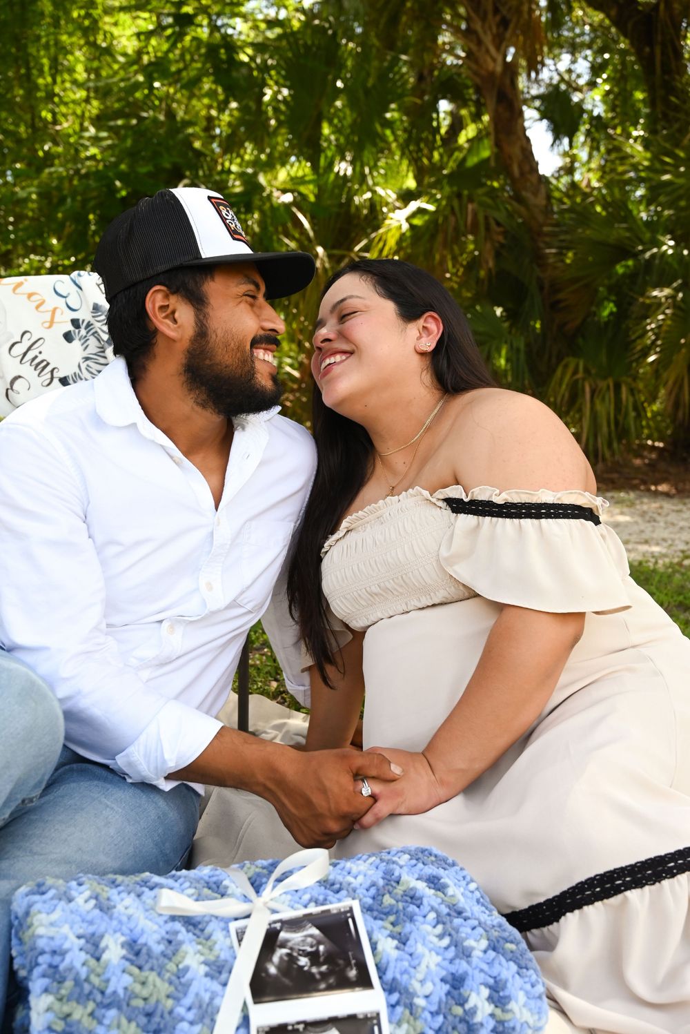 Happy couple sharing a joyful moment outdoors at a maternity photoshoot with ultrasound photos and blue blanket.