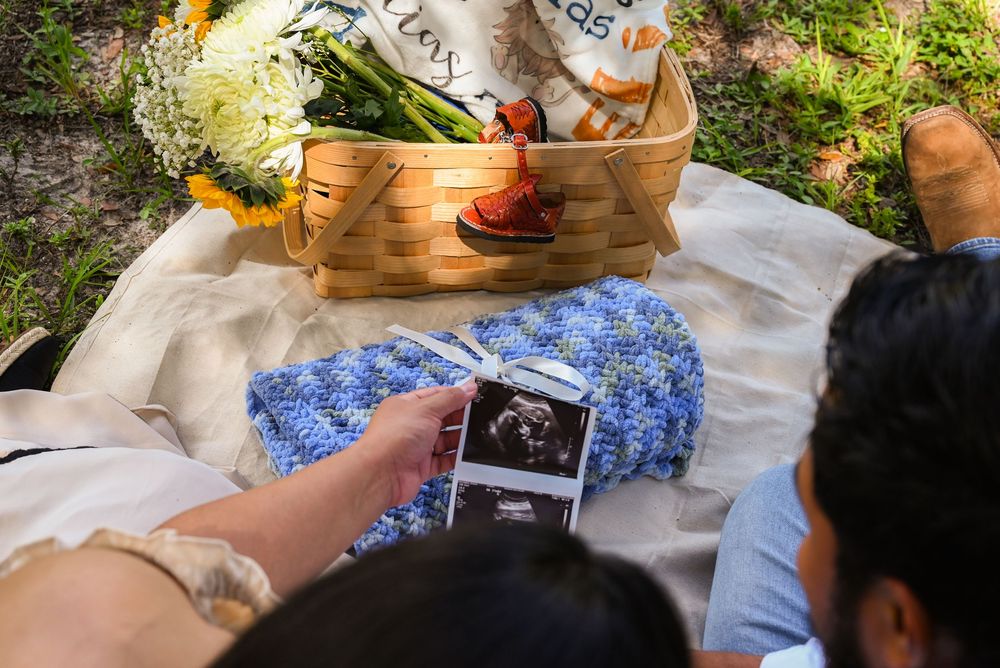 Couple holding ultrasound photos at outdoor picnic with blue knit blanket and wicker basket, pregnancy announcement.