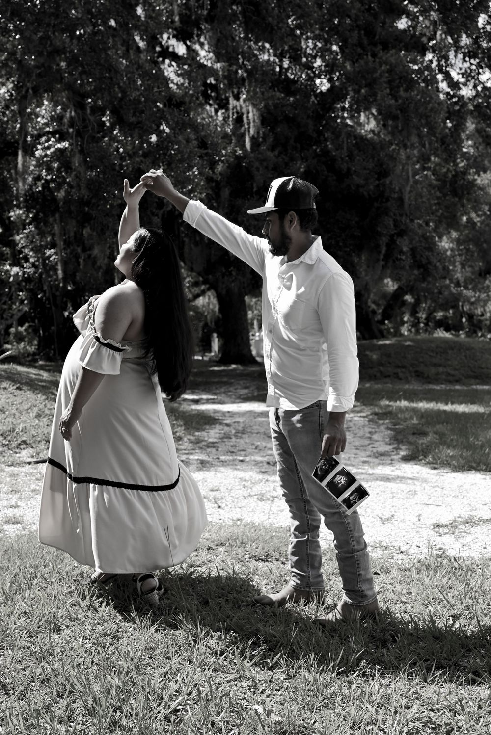 Black and white photo of a couple dancing outdoors, man holding ultrasound photo while spinning pregnant woman in white dress.
