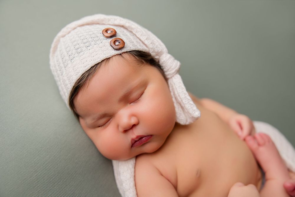 Peaceful newborn baby sleeping on soft sage green blanket wearing white knit hat.