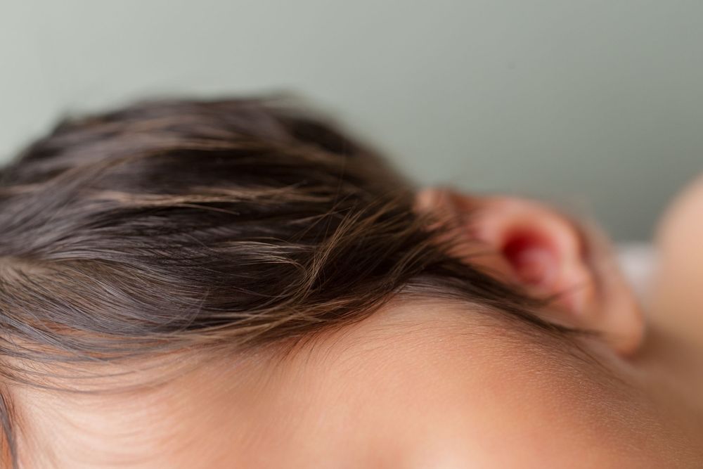 Close-up profile of sleeping newborn with dark hair against sage green backdrop.