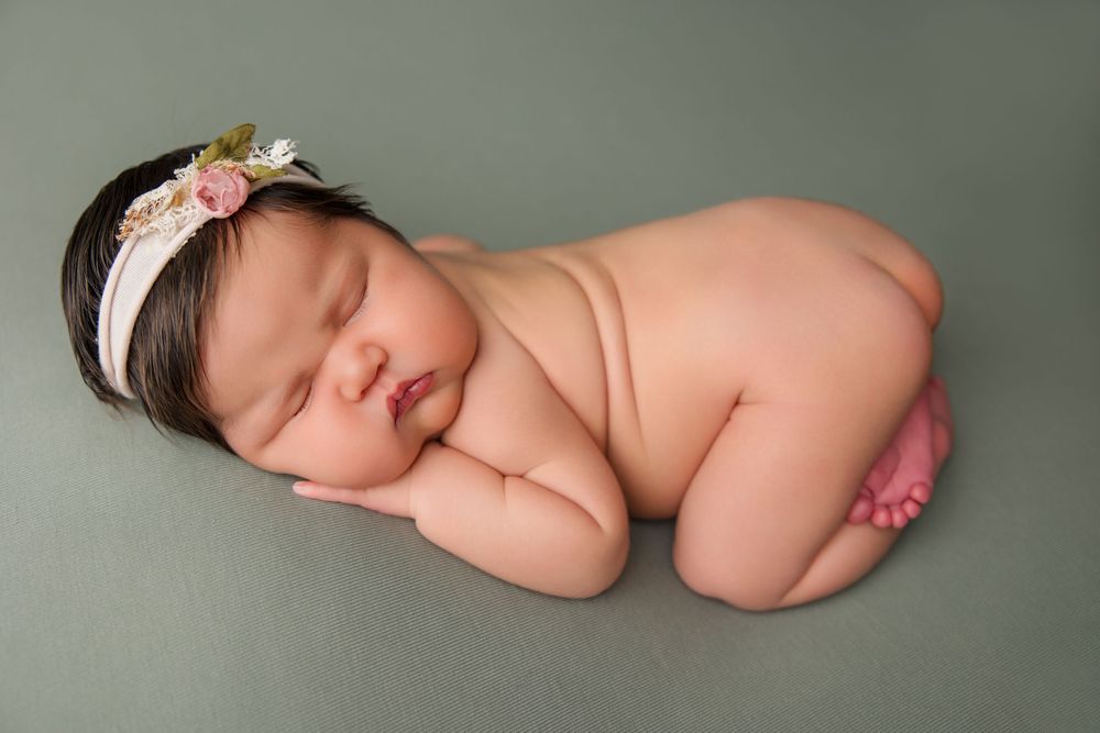 Peaceful sleeping newborn baby curled up on soft sage green backdrop in natural lighting.
