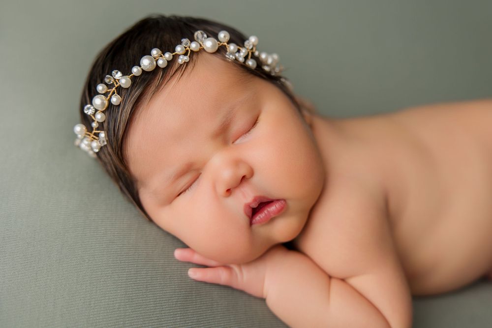 Sleeping newborn wearing delicate crystal headband on sage green background.