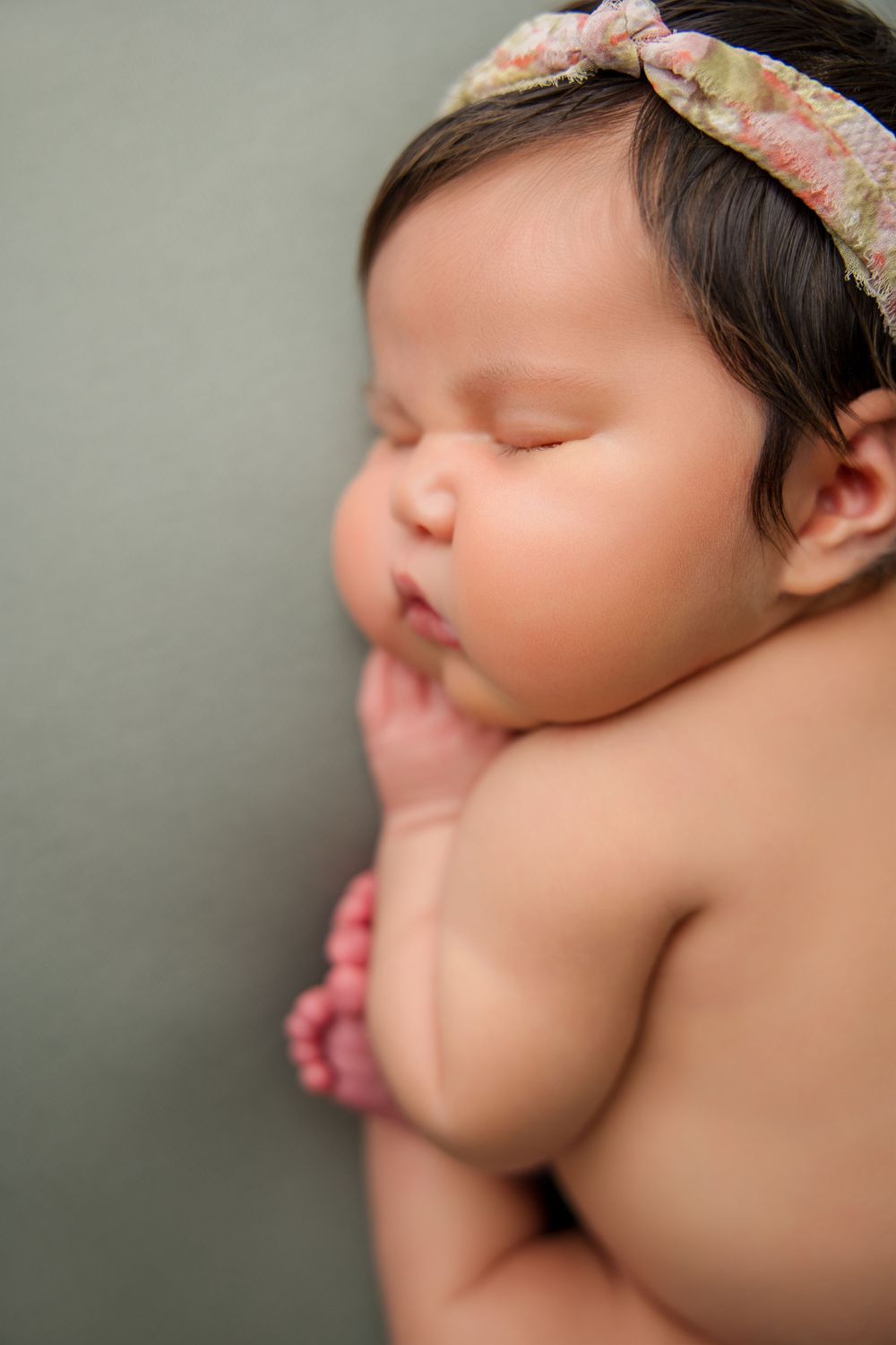 Side profile of a sleeping infant wearing a delicate headband against a gray backdrop.