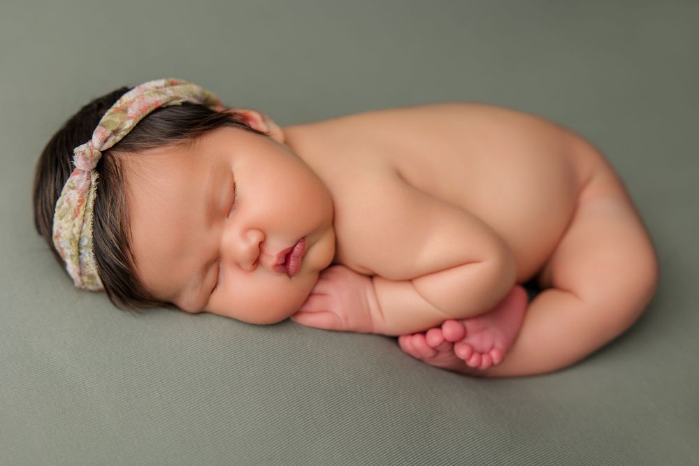 Newborn baby sleeping on muted green background wearing delicate lace headband.