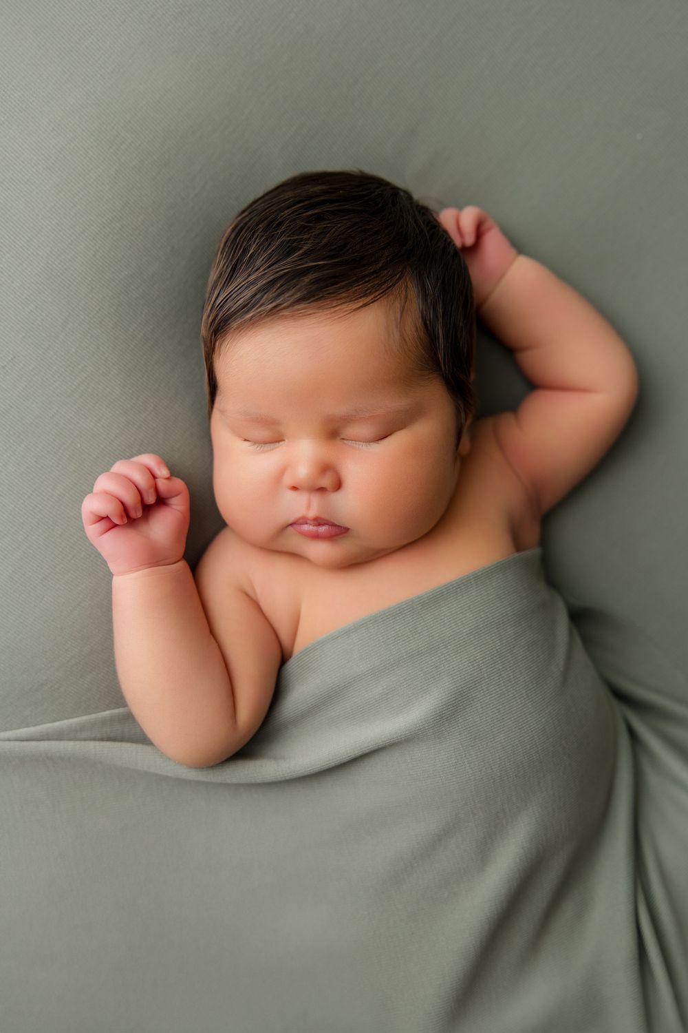 A sleeping newborn baby lies peacefully on soft sage green fabric with tiny hands raised above their head.