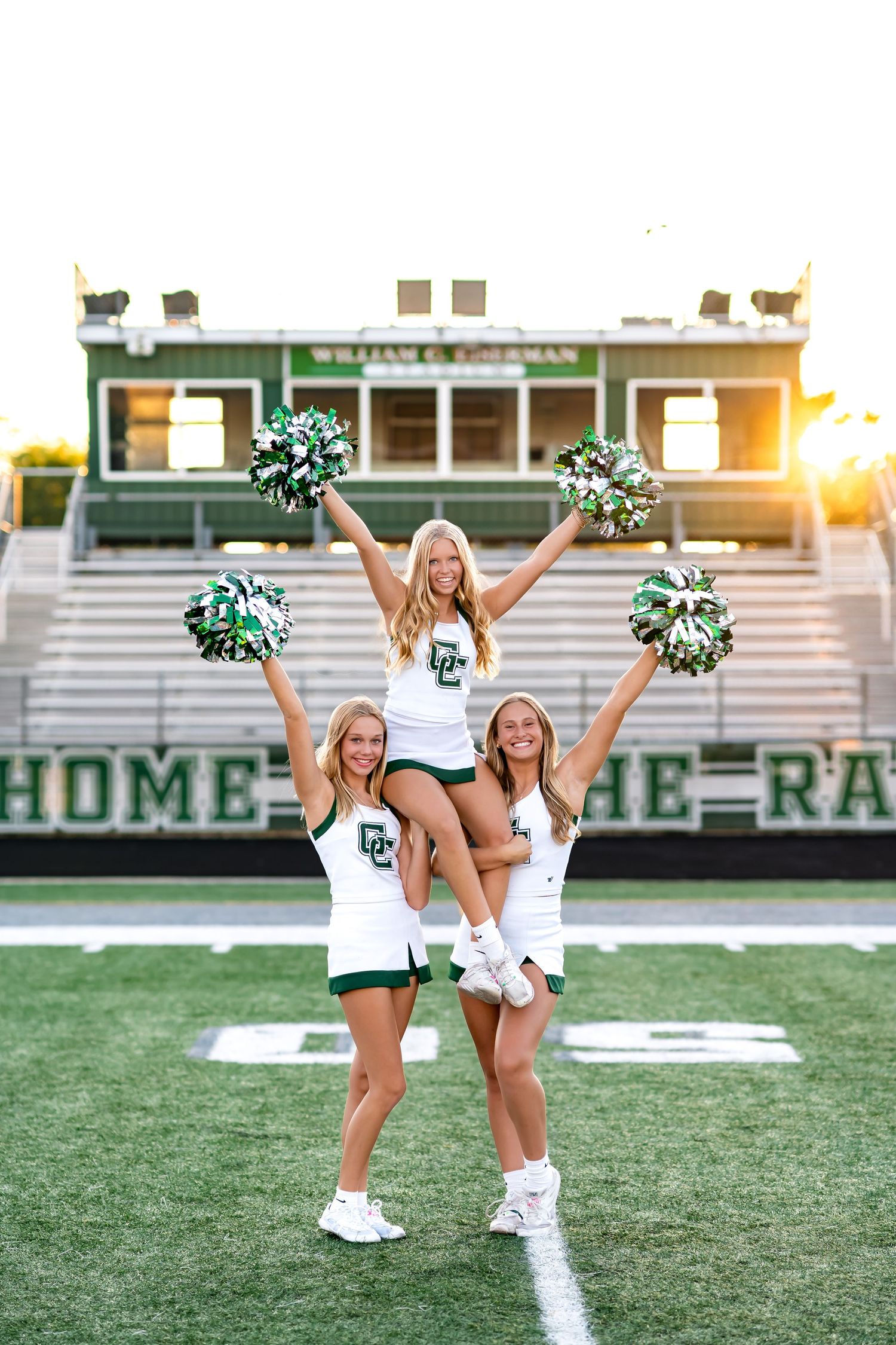 Three cheerleaders in white uniforms perform energetic poses on a football field at sunset with stadium bleachers visible.