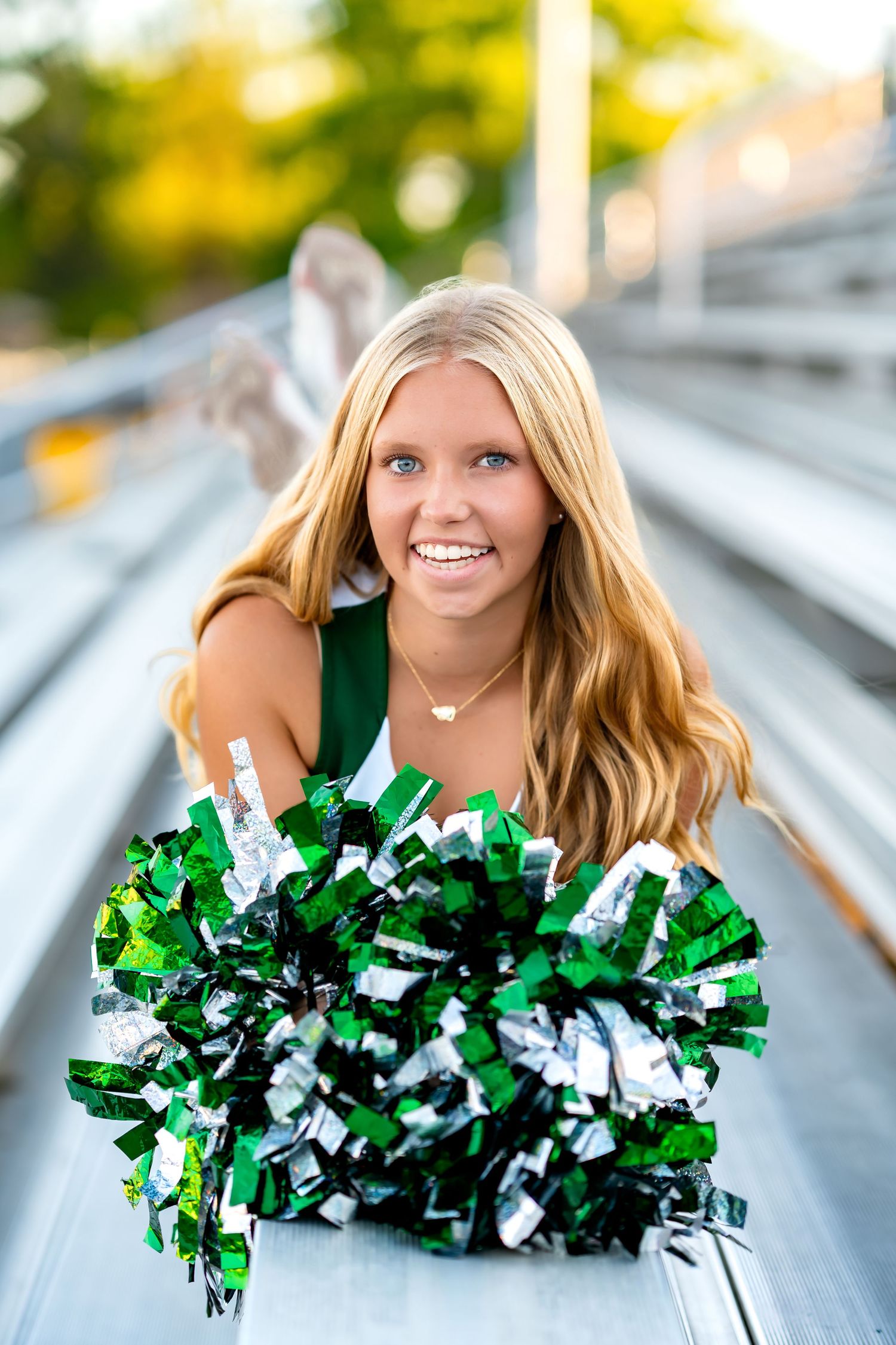 Green and white cheerleading pom poms held up in front of bleachers on a sunny fall day.