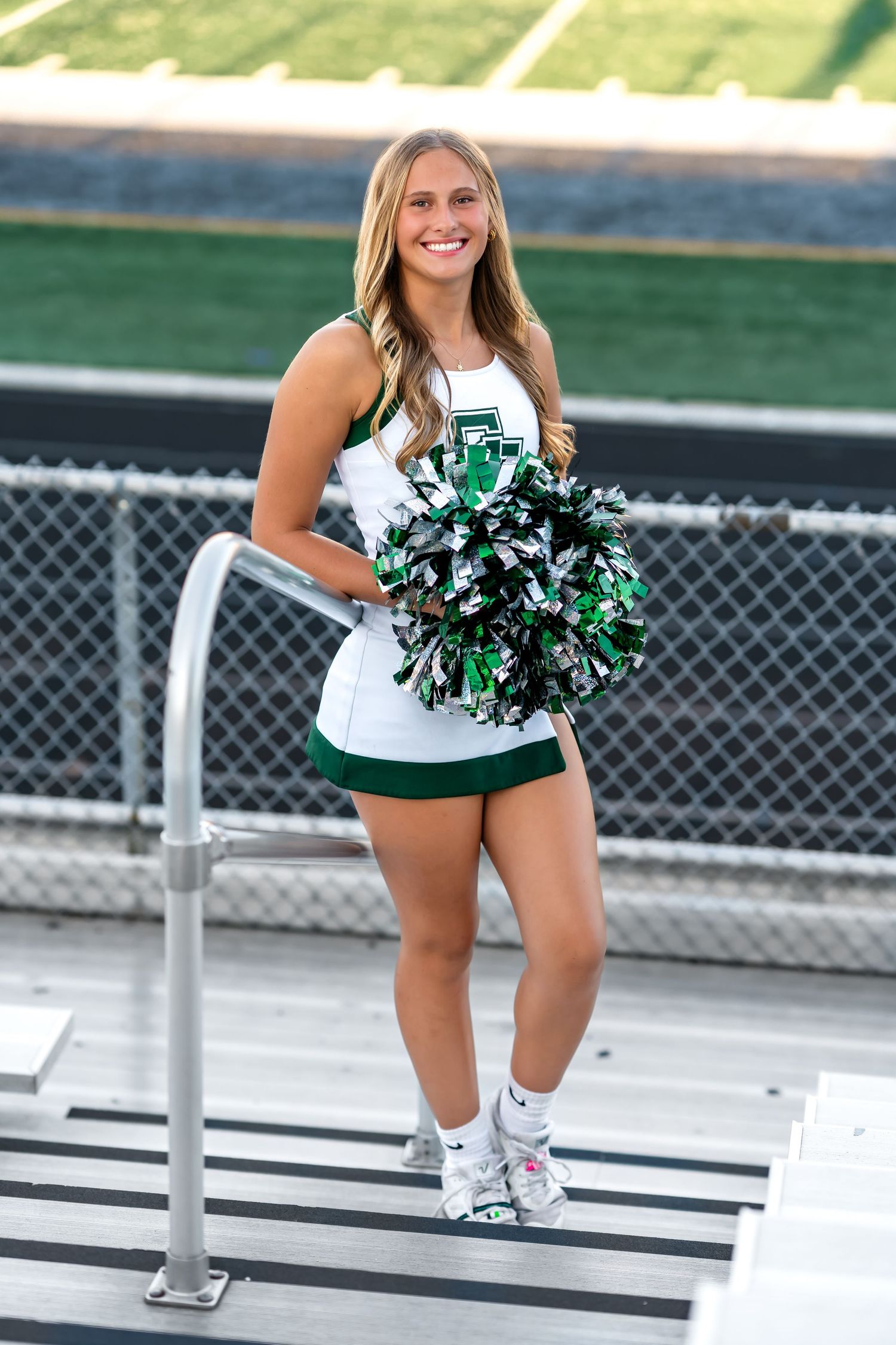 Cheerleader performs a dynamic stance with green and white pom poms on bleacher steps at a sports venue.