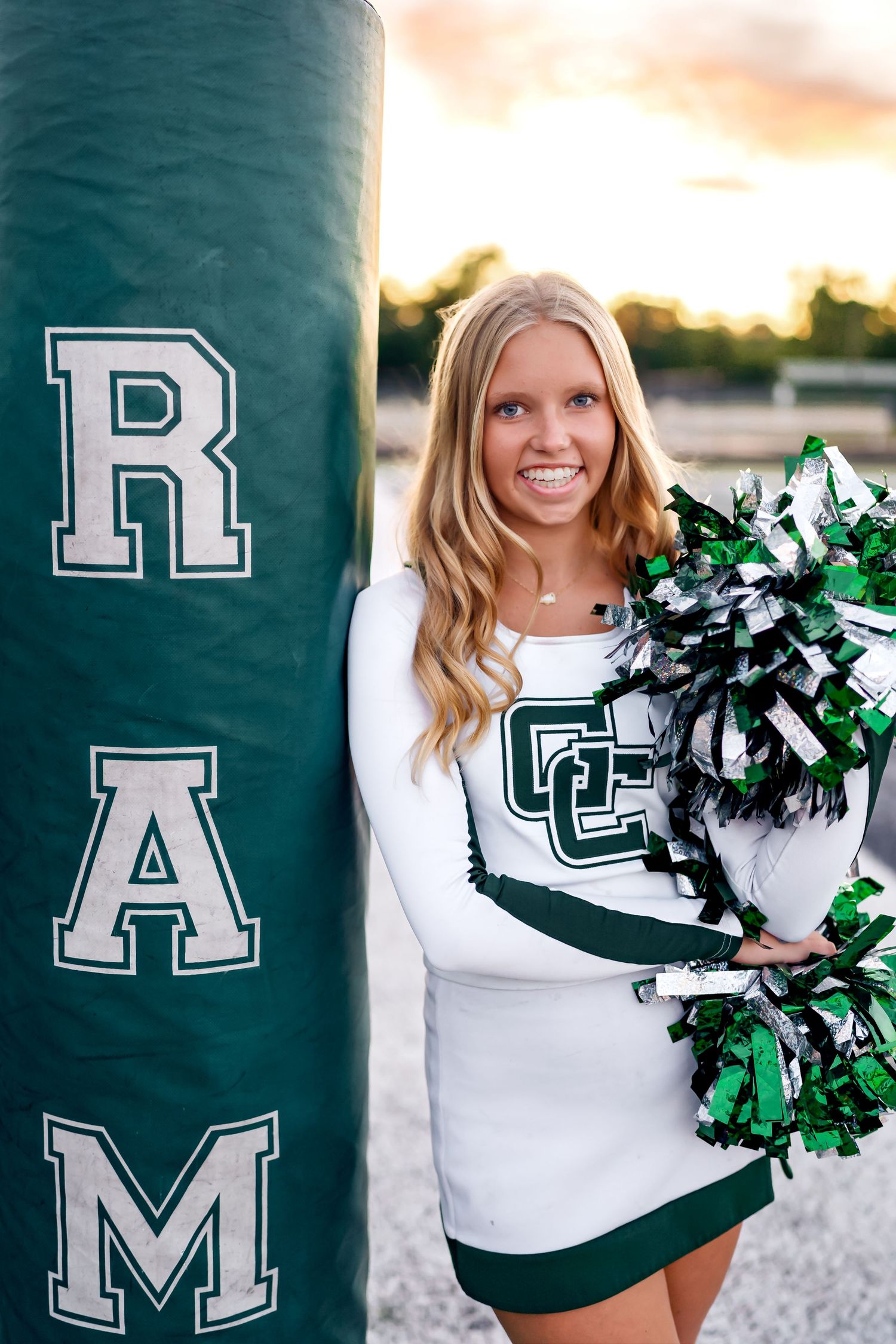 Cheerleader in white and green uniform holds pom poms during outdoor sunset photo session.