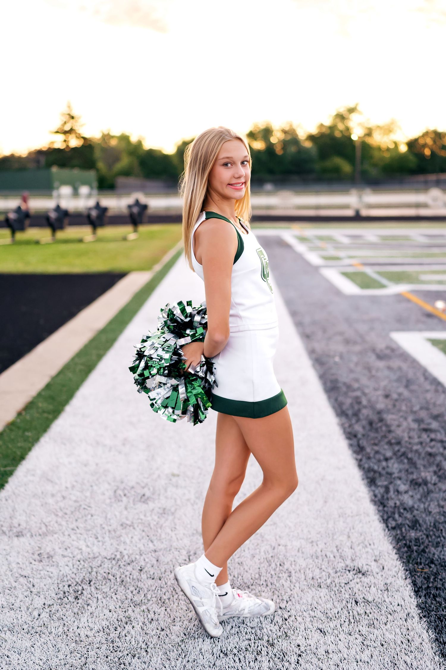 Cheerleader holding green and white pom poms poses in athletic stance on track field at sunset.